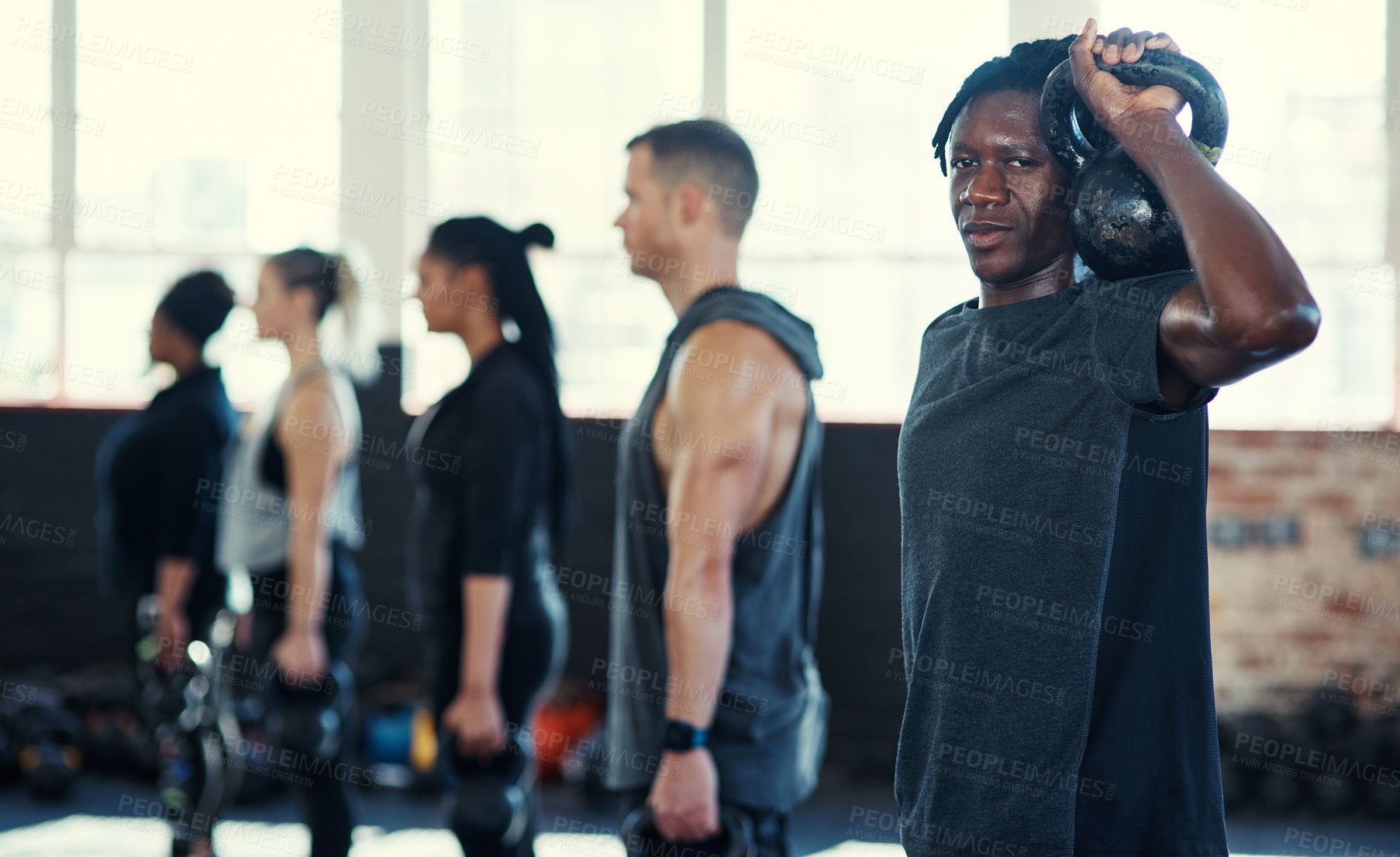 Buy stock photo Shot of a focused group of young people standing in a row and training with weights  while one looks into the camera in a gym