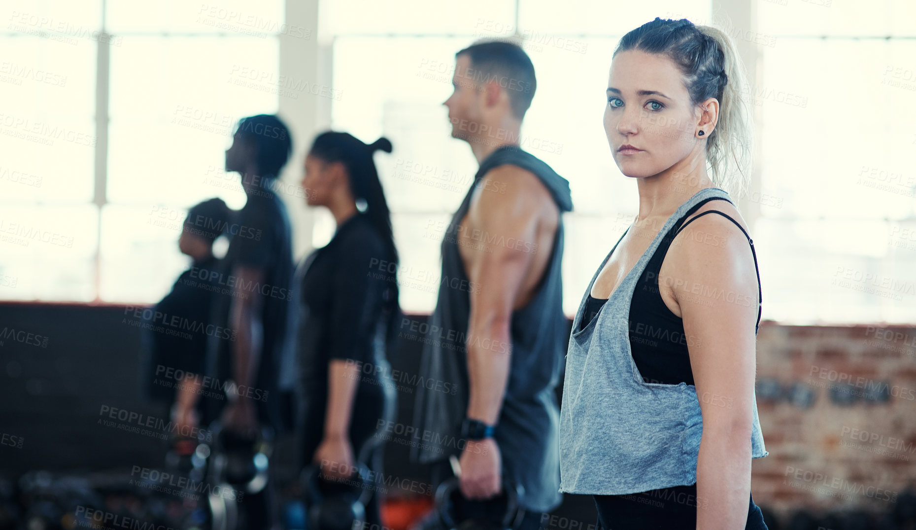 Buy stock photo Shot of a focused group of young people standing in a row and training with weights while one looks into the camera in a gym
