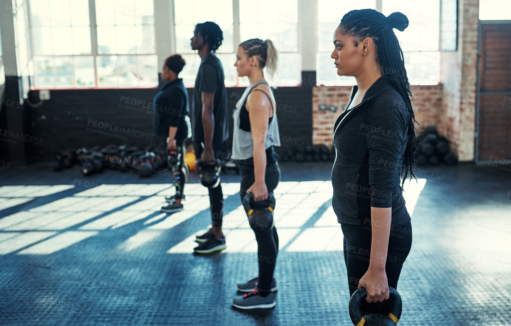 Buy stock photo Shot of a focused group of young people standing in a row and training with weights in a gym