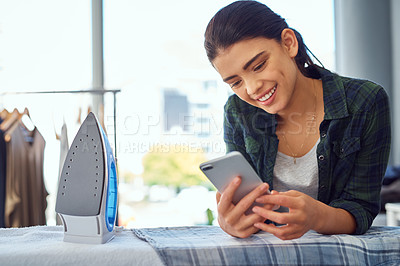 Buy stock photo Cropped shot of an attractive young woman using her cellphone while doing the ironing at home
