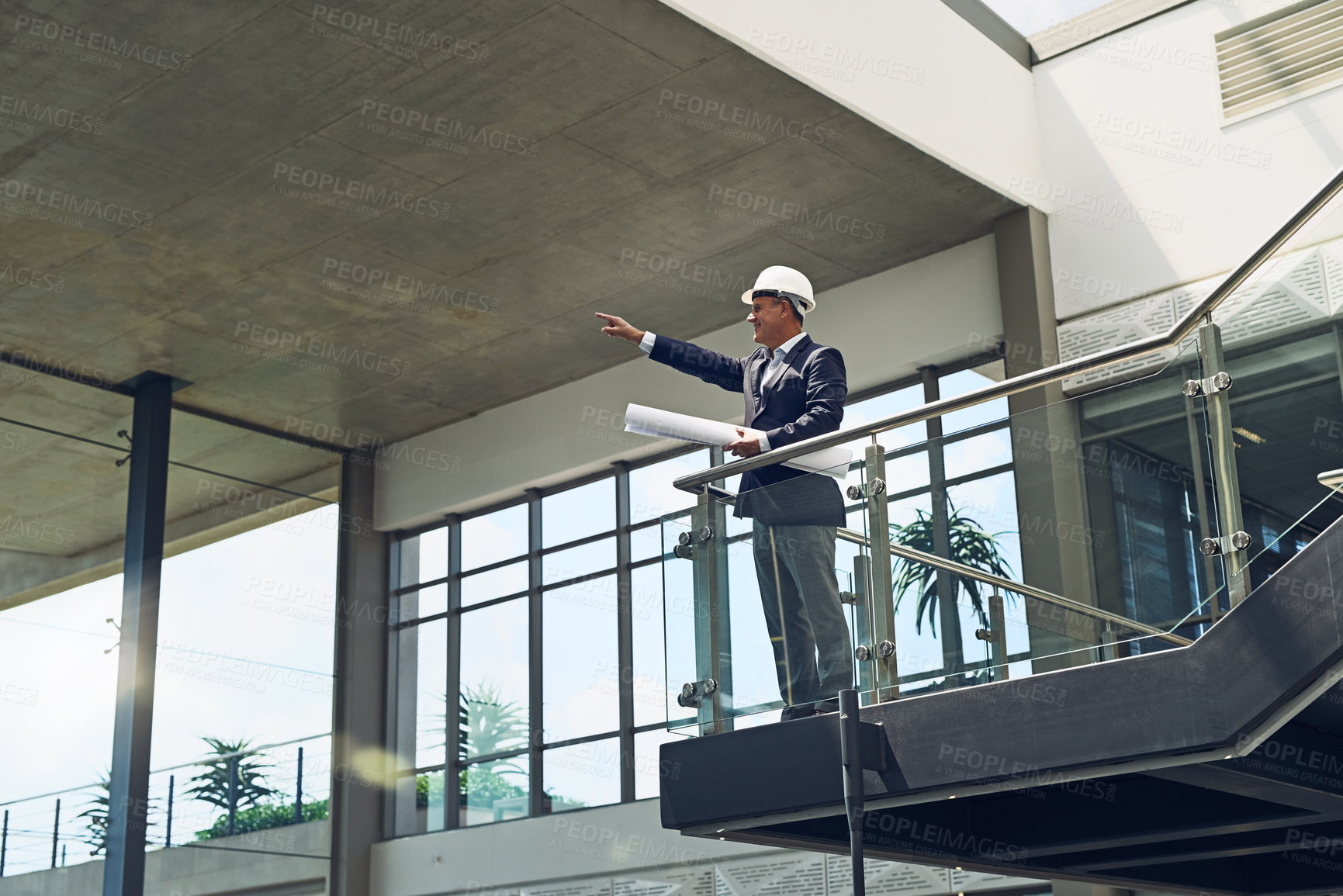 Buy stock photo Shot of a cheerful professional male architect looking at a development site while holding blueprints inside a building