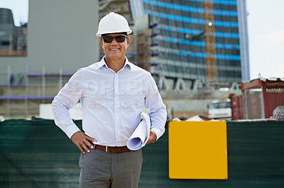 Buy stock photo Portrait of a professional male architect standing on a building site while holding blueprints and looking at the camera outside