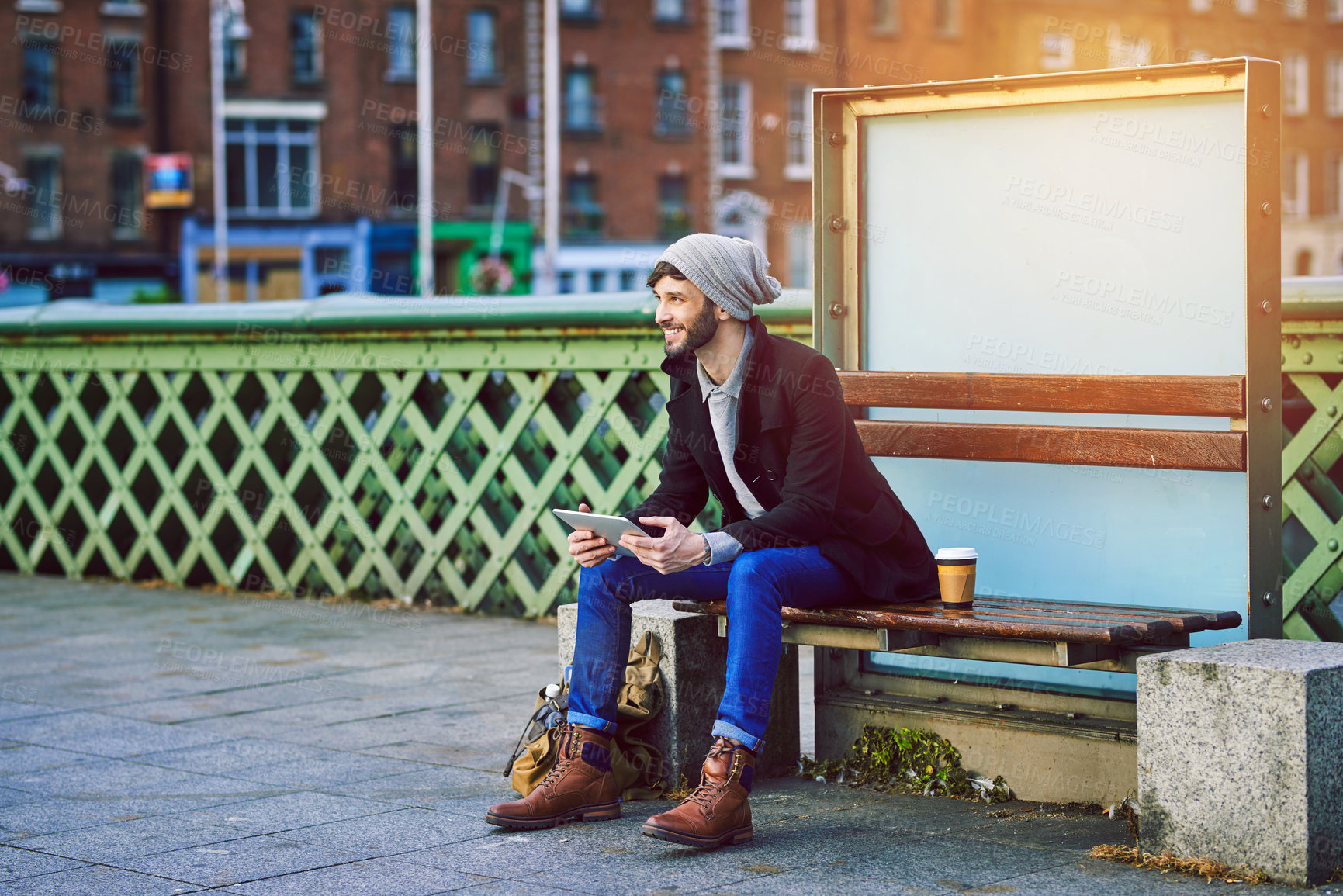 Buy stock photo Shot of a young man using a digital tablet while waiting at a city bus stop