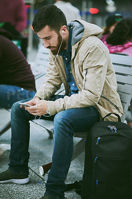 Buy stock photo Full length shot of a handsome young man listening to music on his cellphone while sitting in a bus station