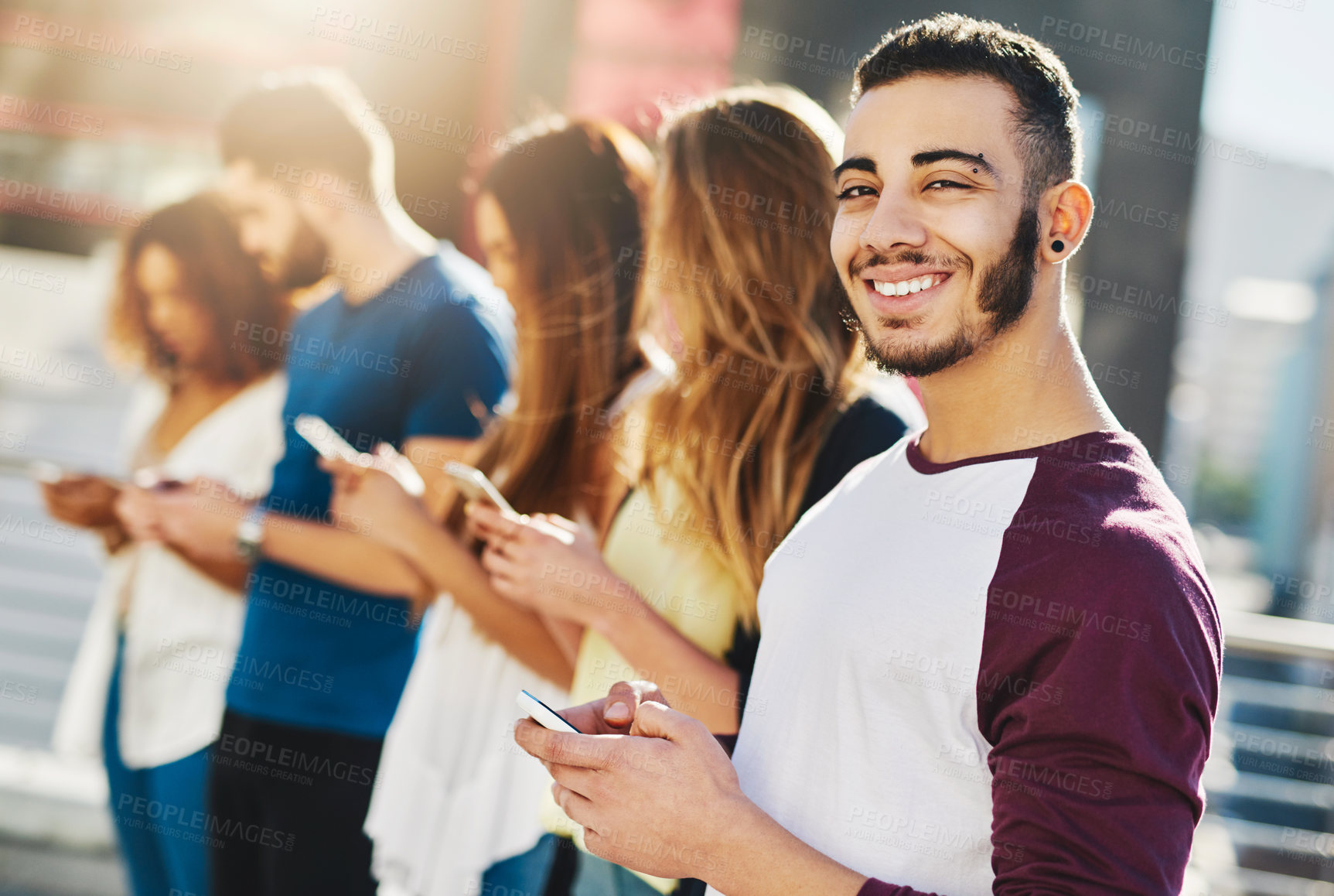 Buy stock photo Cropped portrait of a handsome young man sending a text message while standing outside with a group of friends