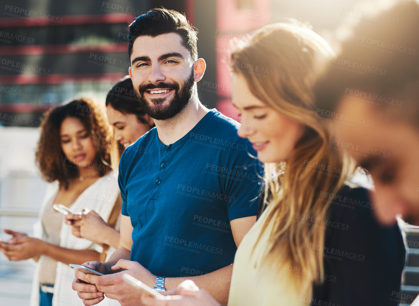 Buy stock photo Cropped portrait of a handsome young man sending a text message while standing outside with a group of friends