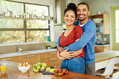Buy stock photo Cropped portrait of a handsome young man embracing his wife while she makes a fruit salad in the kitchen