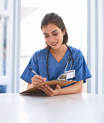 Buy stock photo Shot of a cheerful young female doctor standing while making notes on a clipboard inside a clinic