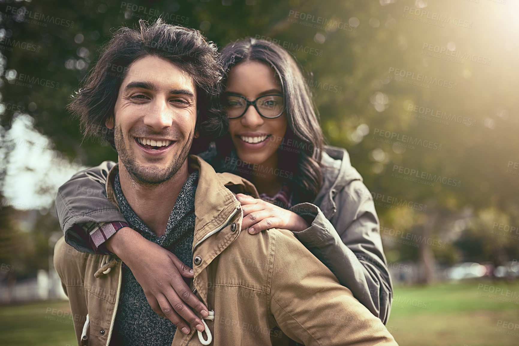Buy stock photo Portrait of a cheerful young couple having a piggyback ride while looking at the camera outside in a park