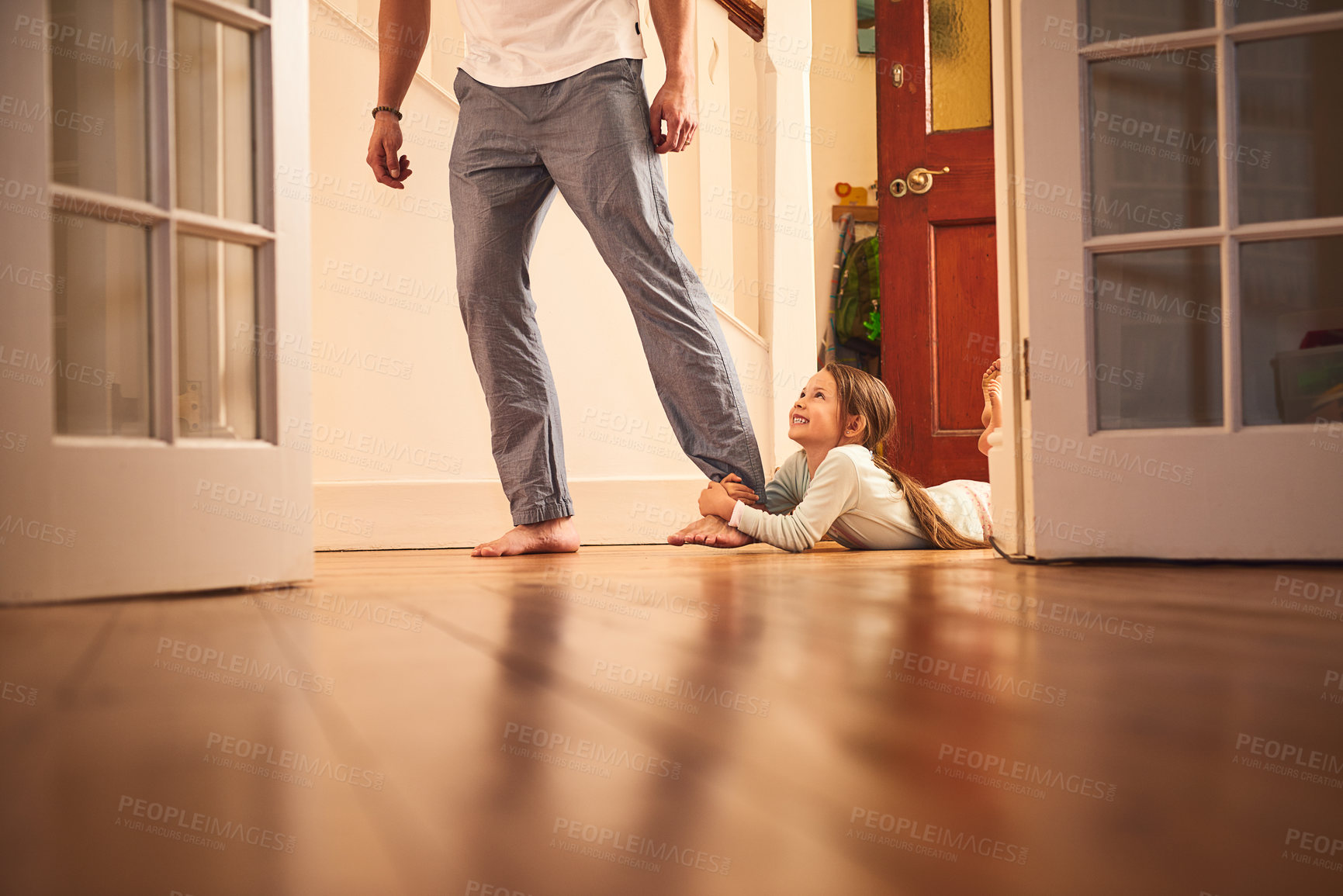 Buy stock photo Shot of a cheerful little girl grabbing hold of her dad's leg and not letting go while lying on the ground at home