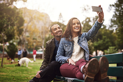 Buy stock photo Shot of a cheerful young couple sitting down on a bench while taking self portraits together outside in a park
