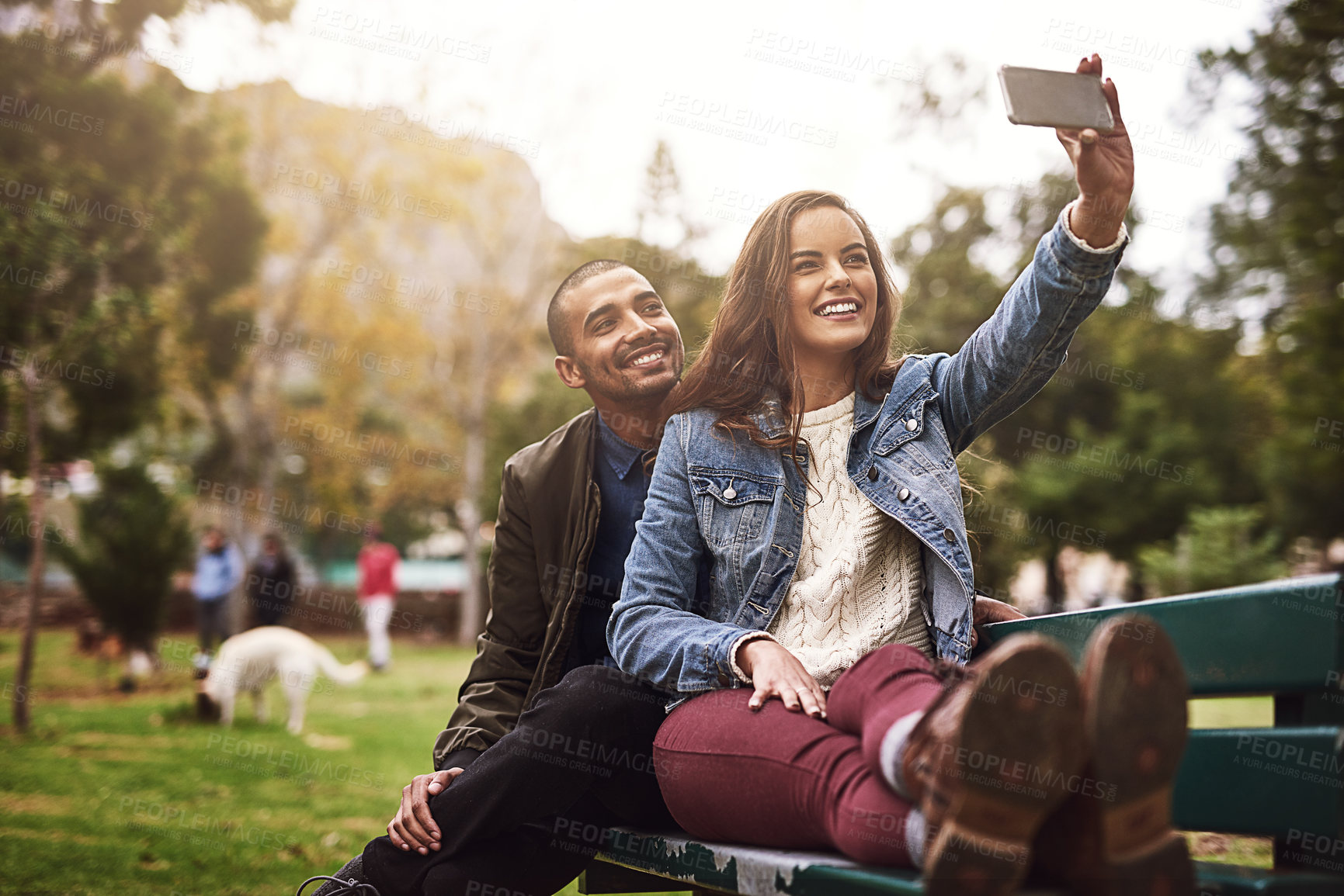 Buy stock photo Shot of a cheerful young couple sitting down on a bench while taking self portraits together outside in a park