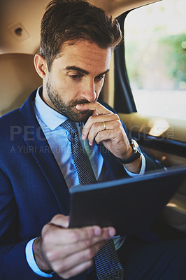 Buy stock photo Shot of a focused young businessman working on his digital tablet while being seated in the back of a car