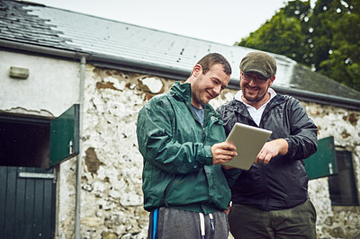 Buy stock photo Shot of two cheerful young farmers busy on a digital tablet while standing outside on a farm during the day