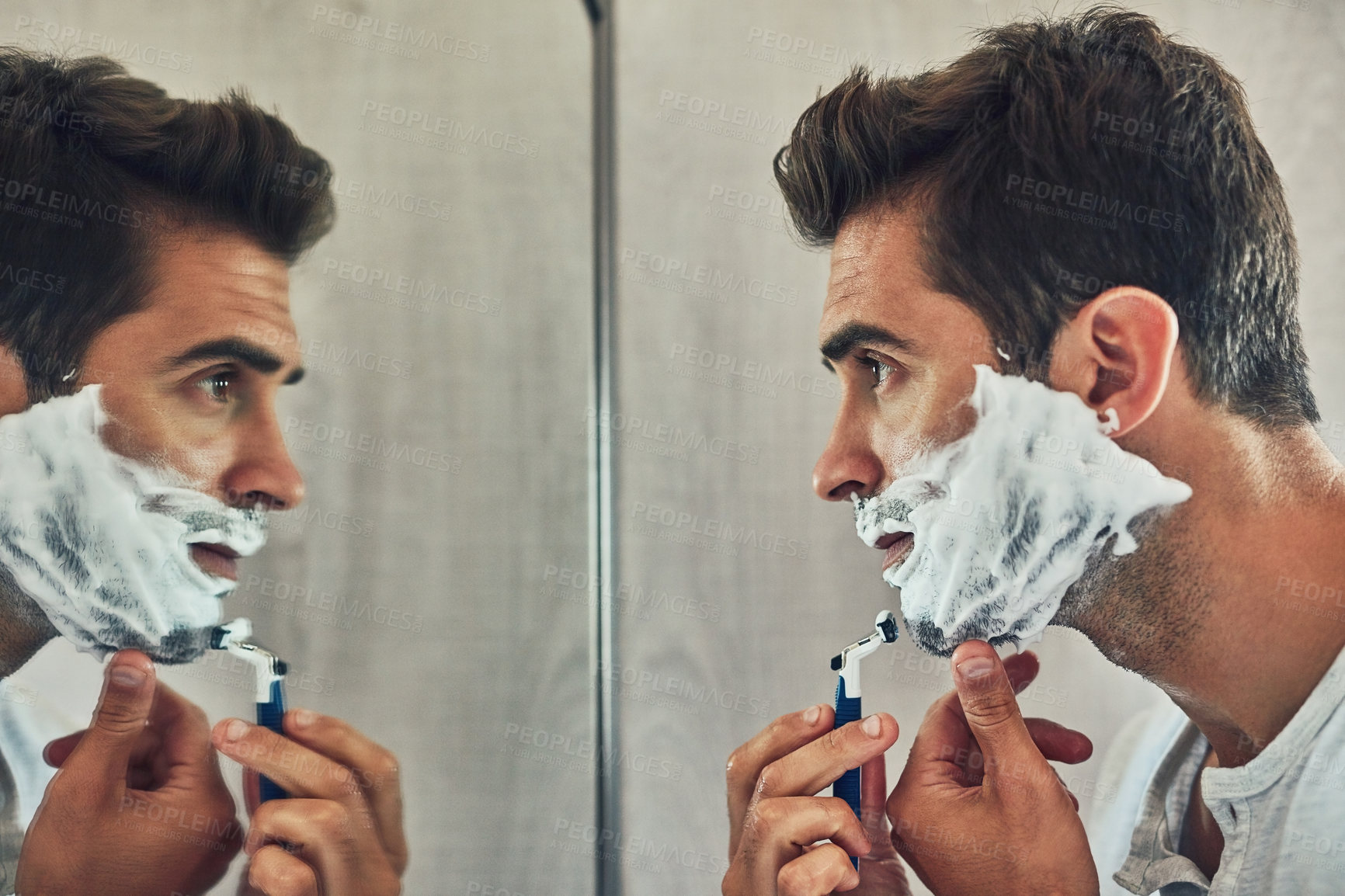 Buy stock photo Shot of a focused young man shaving his beard while looking at his reflection in the mirror at home