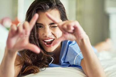 Buy stock photo Portrait of an attractive young woman looking at you through a finger frame while relaxing in her bedroom
