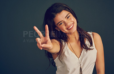Buy stock photo Studio shot of a cheerful young woman showing the peace sign while standing against a dark background