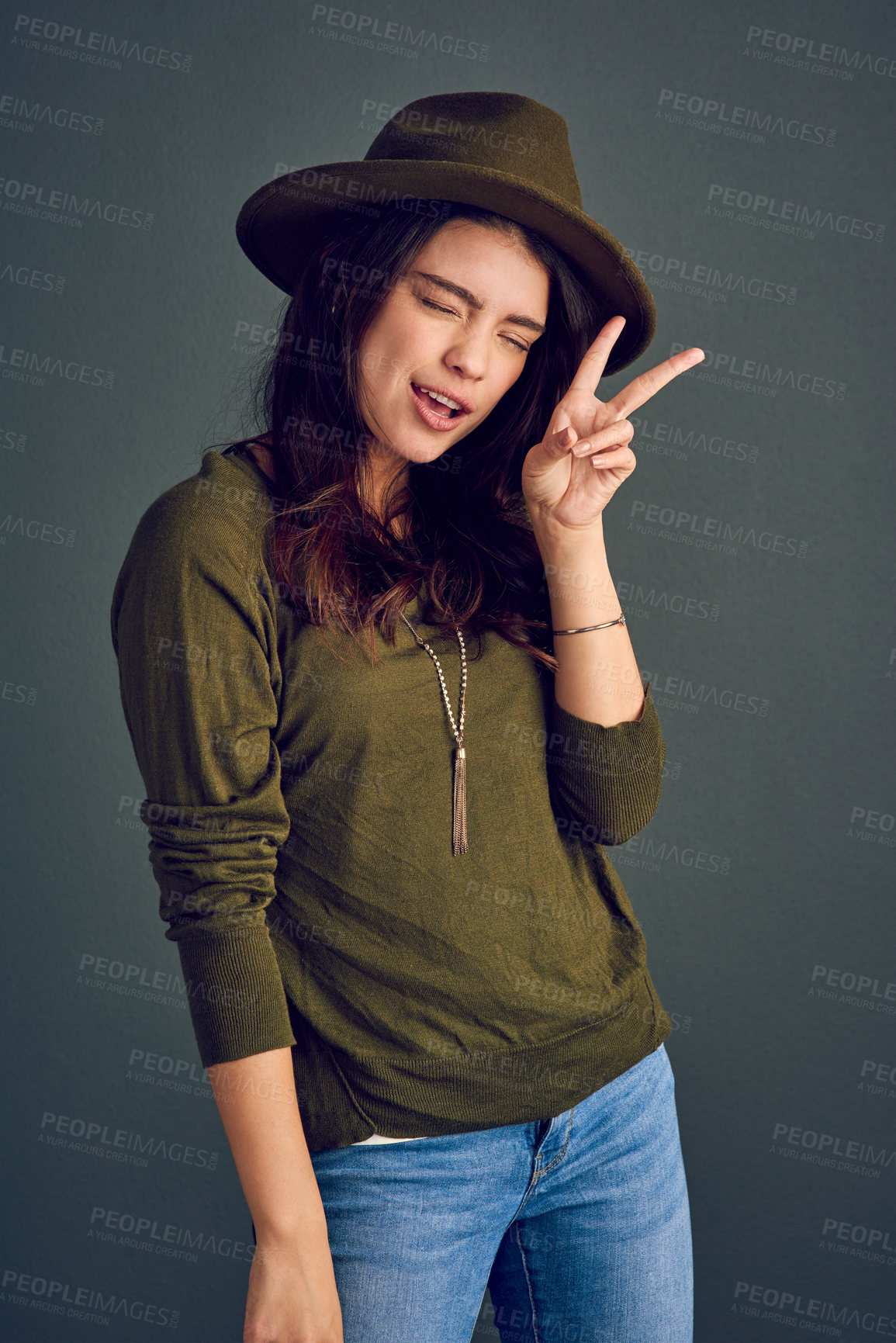 Buy stock photo Studio shot of a carefree young woman posing with a hat and showing the peace sign while standing against a dark background