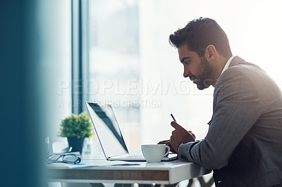 Buy stock photo Shot of a handsome young businessman using a laptop and cellphone at his desk in a modern office
