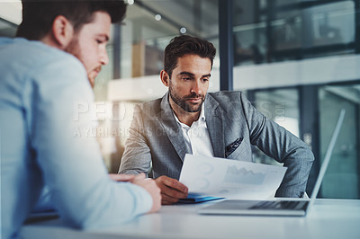 Buy stock photo Shot of two young businessmen using a laptop while going through paperwork together in a modern office