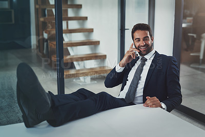 Buy stock photo Shot of a handsome young businessman relaxing with his feet up on an office desk while using his phone
