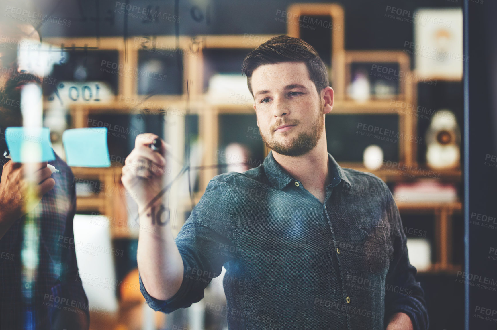 Buy stock photo Shot of two young businessmen brainstorming on a glass wall in an office