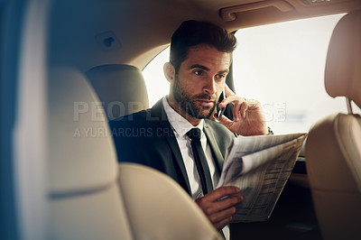 Buy stock photo Shot of a confident young businessman talking on his phone and looking at a newspaper in the backseat of a car