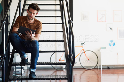 Buy stock photo Shot of a young designer working on his tablet while sitting on a staircase in the office