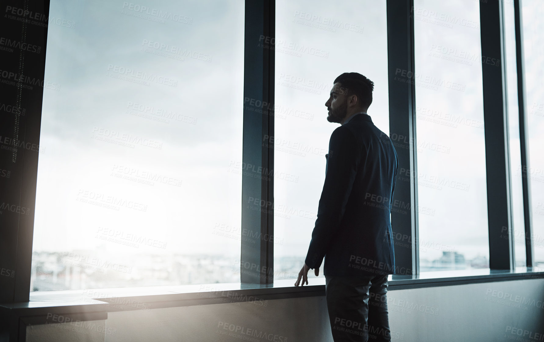 Buy stock photo Shot of a young businessman looking thoughtfully out of an office window