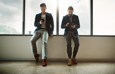 Buy stock photo Shot of two young businessmen using a digital tablet and cellphone while leaning against a wall in a modern office