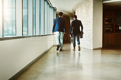 Buy stock photo Rearview shot of two young businessmen walking down a corridor of a modern office