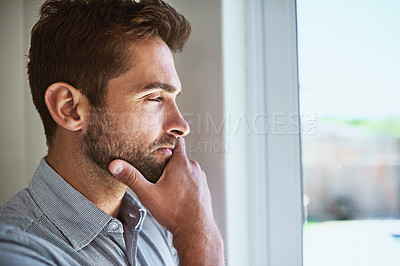Buy stock photo Shot of a focused young man looking through a window while contemplating inside at home during the day