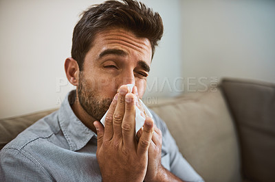 Buy stock photo Portrait of a uncomfortable looking young man sneezing into a tissue while being seated on a couch at home