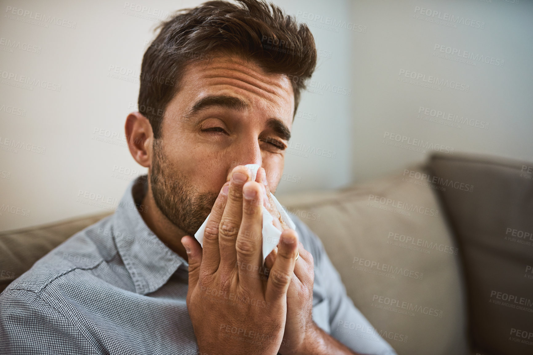 Buy stock photo Portrait of a uncomfortable looking young man sneezing into a tissue while being seated on a couch at home