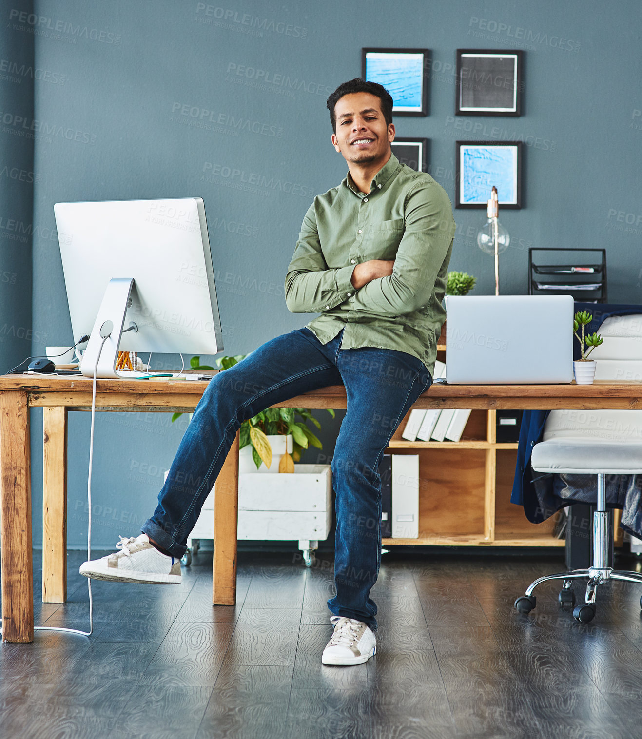 Buy stock photo Portrait of a cheerful young businessman seated on his desk with his arms folded inside of the office during the day