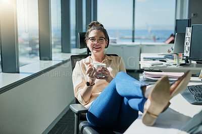 Buy stock photo Shot of a cheerful young businesswoman drinking coffee while sitting with her feet up on her desk inside of the office