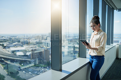 Buy stock photo Shot of a focused young businesswoman browsing on a digital tablet while standing next to a window inside of the office