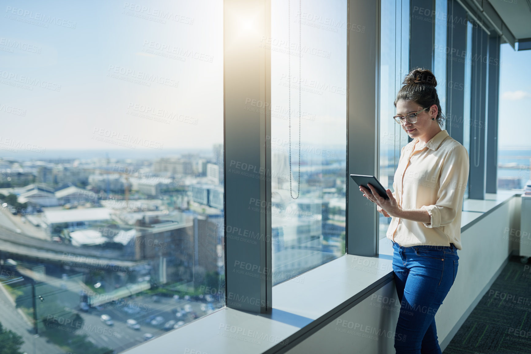Buy stock photo Shot of a focused young businesswoman browsing on a digital tablet while standing next to a window inside of the office