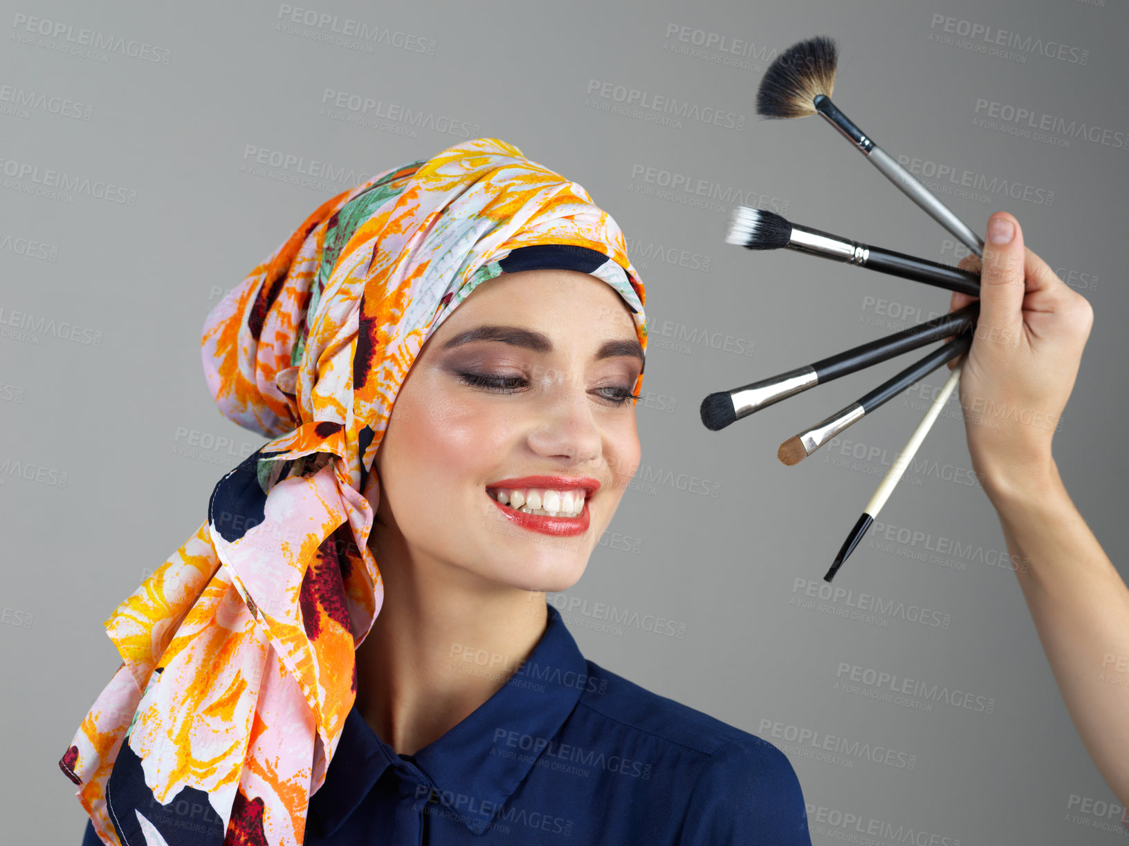 Buy stock photo Studio shot of a confident young woman wearing a colorful head scarf while looking at different makeup brushes being held in front of her