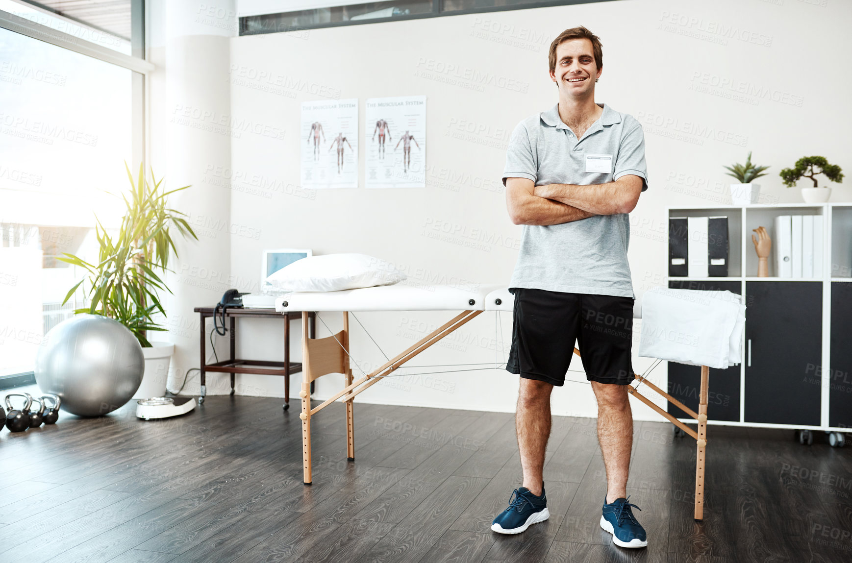 Buy stock photo Portrait of a confident young male physiotherapist standing with his arms folded inside of his office