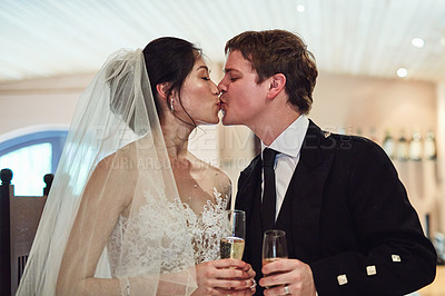 Buy stock photo Shot of a cheerful young bride and groom sharing a kiss with each holding a champagne glass inside of a building