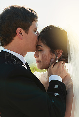 Buy stock photo Shot of a cheerful young groom giving his bride a kiss on the forehead while they stand holding each other outside