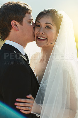 Buy stock photo Portrait of a cheerful young groom giving his bride a kiss on the cheek while they stand holding each other outside