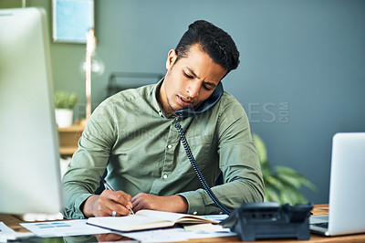 Buy stock photo Shot of a focused young businessman seated at his desk while taking on the phone and making notes inside the office