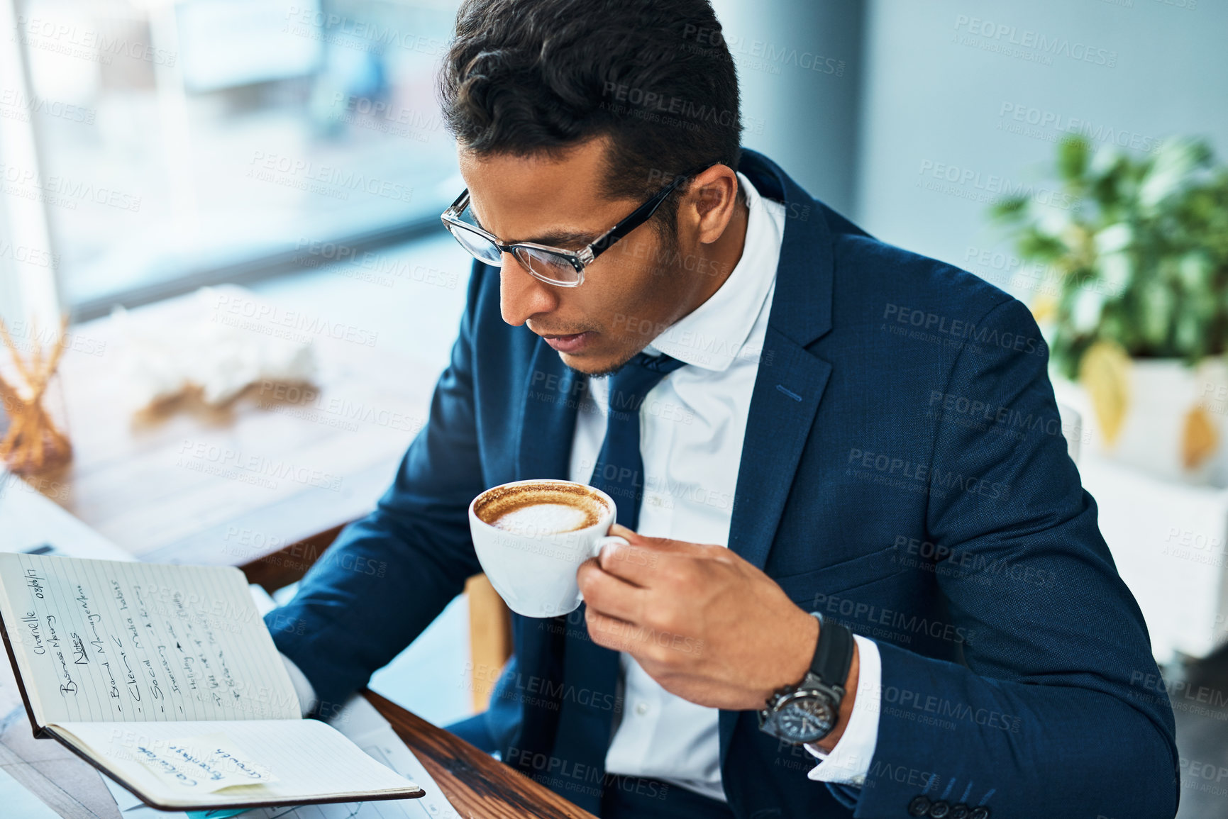 Buy stock photo Shot of a focused young businessman seated at his desk  while drinking coffee and reading his journal in the office