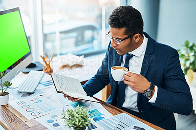 Buy stock photo Shot of a focused young businessman seated at his desk  while drinking coffee and reading his journal in the office