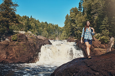 Buy stock photo Shot of an attractive young woman walking next to a rocky river and waterfall