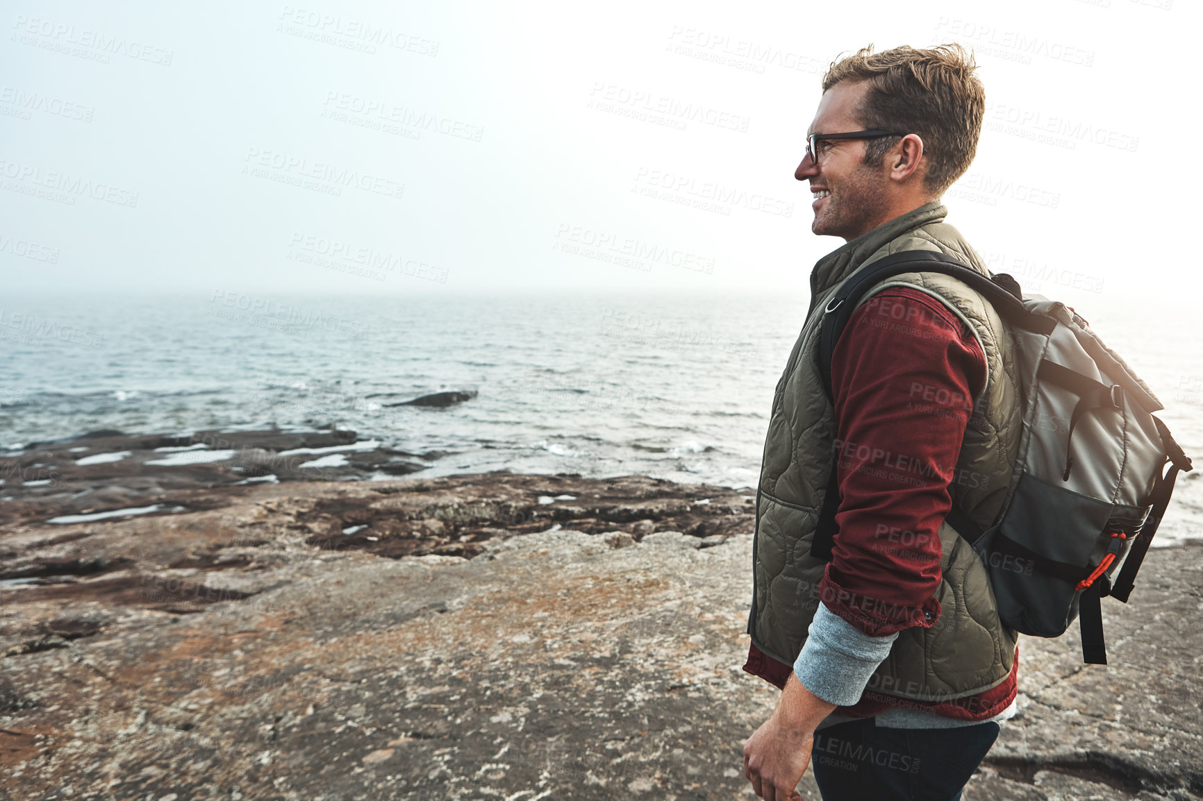 Buy stock photo Shot of a cheerful middle aged man walking on rocks next to the ocean while wearing a backpack outside during the day