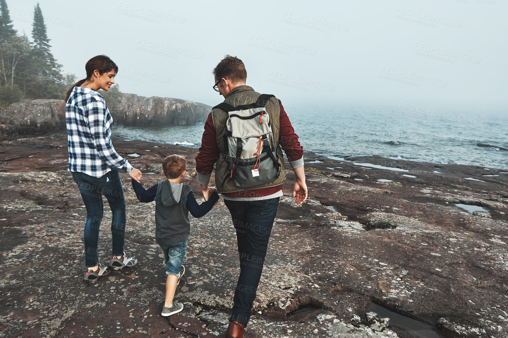 Buy stock photo Rearview shot of a cheerful young family holding hands and walking together next to the ocean outside during the day