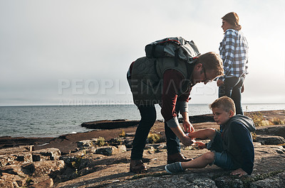 Buy stock photo Shot of a cheerful man helping to put his son's shoes on while standing next to the ocean outside during the day